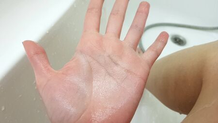 Wrinkled female hand because of soak in the water for a long time, wet white bathroom and leg on...