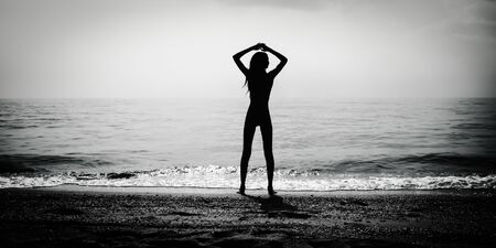 Dark silhouette of slim girl on the sea beach with hands by head, bw photoの写真素材
