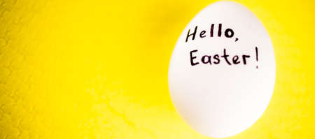 White chicken egg with black inscription hello easter on a yellow backgroundの写真素材