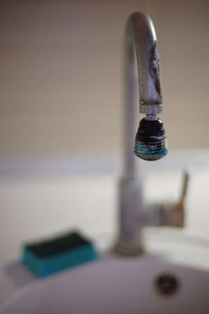 The old chrome faucet in the kitchen sink is covered in limescale. Blue dish sponge lies on a white sink.の写真素材