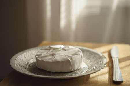 Whole round Camembert cheese on a silver plate with a knife on wooden table.