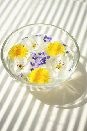 White blooming flowers, yellow dandelions and purple seeds in a glass bowl on a white sunny table.