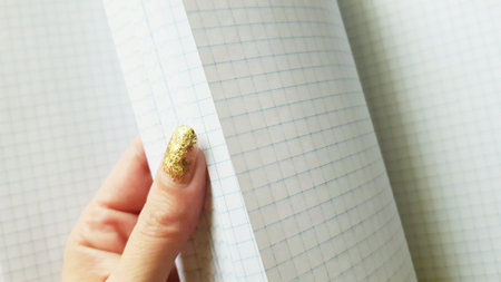 A beautiful female hand with a golden manicure flips through a blank checkered notebookの写真素材