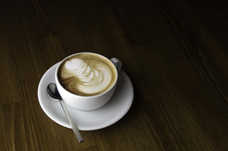 coffee with milk in a white cup on a brown table and spoon to mixの写真素材