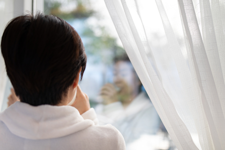 Rear view of Asian woman drinking a glass of milk in living room and looking outside window in the morningの写真素材