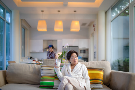 Asian woman sitting on sofa in living room with a glass of milk in hand and mom standing in backgroundの写真素材