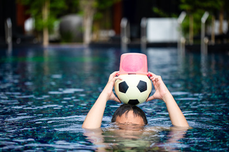 Asian boy put a ball and plastic bucket on head in swimming poolの写真素材