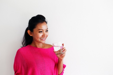 Young woman in a pink top drinking water and standing against a white wallの写真素材