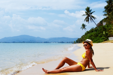Young woman in a yellow swimsuit, a hat and sunglasses seating down on a sandy beach of Thailand and sun bathing on a sunny summer dayの写真素材