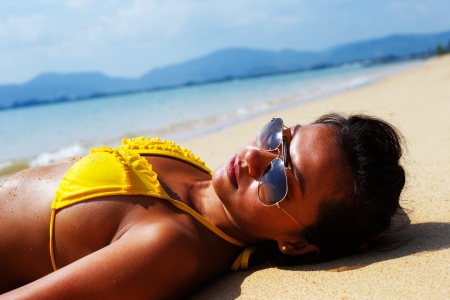 Young woman in a yellow swimsuit and sunglasses laying down on a sandy beach of Thailand on a sunny summer dayの写真素材