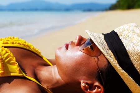 Young woman in a yellow swimsuit, a hat and sunglasses laying down on a sandy beach of Thailand on a sunny summer dayの写真素材