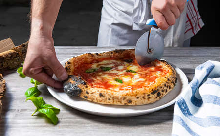 The chef is slicing a fresh pizza. Margarita Pizza straight from the wood-fired oven. Close-up, baked in a wood-fired pizza oven. Italian cuisine. Rustic style. Culinary and restaurant concept.の写真素材
