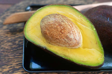 Freshly sliced Hass avocado halves lying on a black tray and a chopping board made of palm tree....