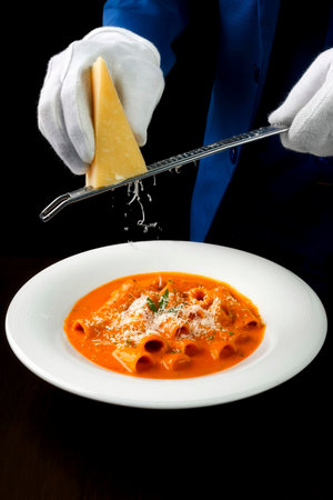 Chef grating Parmesan over rigatoni pasta in creamy tomato sauce with fresh herb garnish. Italian cuisine. Isolated on white.の写真素材