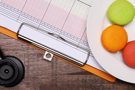 a doctor's stethoscope and a piece of patient's vital sign sheet with a plate of macarons on top of a natural brown table topの写真素材