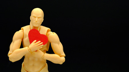 a plastic man model holding a piece of red heart made of wood, isolated on black background