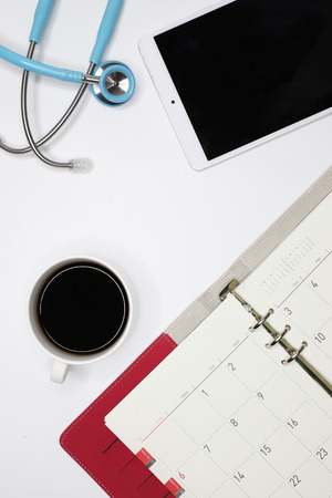 a top view image of a doctor's clean desk with an appointment book, a tablet, a cup of coffee, and a stethoscopeの写真素材
