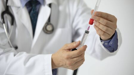 A doctor in white coat drawing a sample from a blood tube with a syringe.