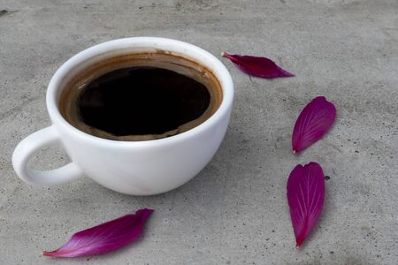 A white ceramic cup of coffee on a grey concrete table  with violet red petals of orchid tree flower. Side view.の写真素材
