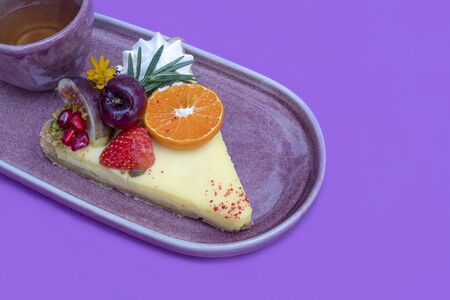 A serving of lemon tart topped with mixed fresh fruit with a cup of green tea on a purple ceramic...