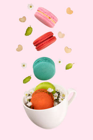 Colorful macaroons, white little flowers, green mint leaf, and wooden hearts levitating on a white tea cup.  Isolation on soft pink background.の写真素材