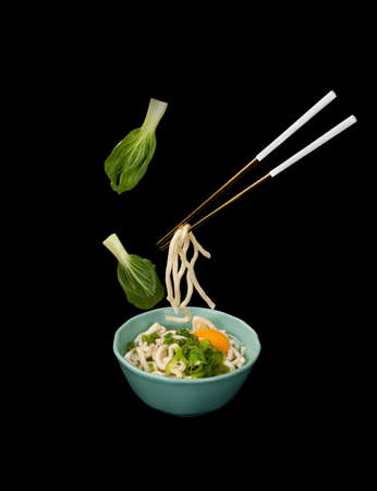Japanese noodles or U-don in a pair of golden chopsticks levitating over a teal ceramic bowl. Levitation food concept.の写真素材