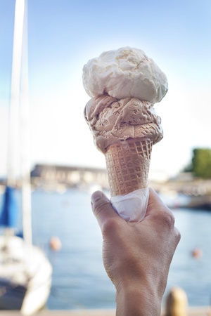 Hand holding an ice-cream cone in front of the harbour on a sunny day 