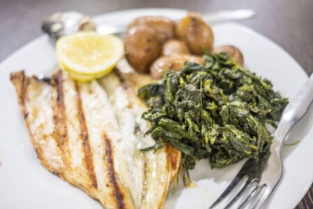 Barbecued sea bass served with lemon, baked potatoes and spinach on white plateの写真素材