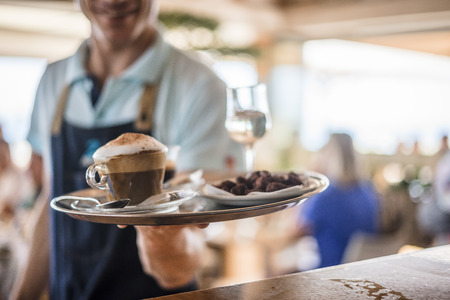 Waiter carries tray with cappuccino, wine glass and desserの写真素材