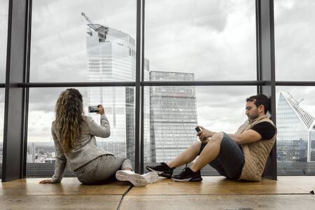 Beautiful young woman taking photo of skyscrapers, while her man is playing with his phone, London, UK. Modern date - together, but separately.の写真素材