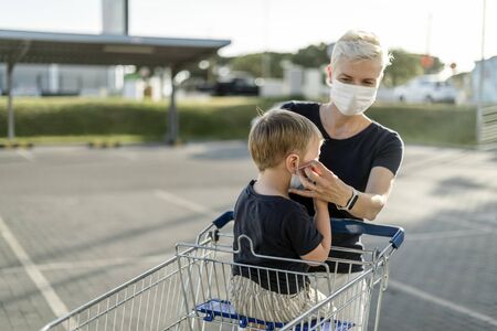 Mother helping her son sitting in a shopping car to wear a protective mask before getting inside a shop.の写真素材