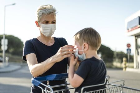 Mother helping her son sitting in a shopping car to wear a protective mask before getting inside a shop.の写真素材