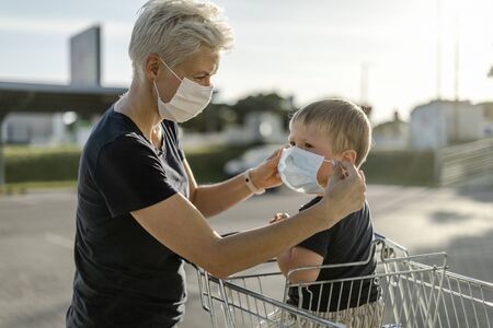 Mother helping her son sitting in a shopping car to wear a protective mask before getting inside a shop.の写真素材