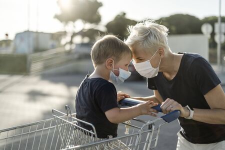 Mother joyfully playing with kid sitting in shopping cart. Both wearing protective face mask before entering a supermarket.の写真素材