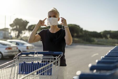 Woman with shopping cart putting protective face mask on just before entering a shopの写真素材