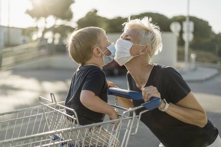 Mother joyfully playing with kid sitting in shopping cart. Both wearing protective face mask before entering a supermarket.の写真素材