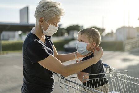 Mother helping her son sitting in a shopping car to wear a protective mask before getting inside a shop.の写真素材
