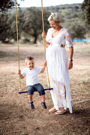 Pregnant woman having fun with her toddler son on the swing in the parkの写真素材