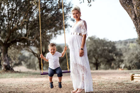Pregnant woman having fun with her toddler son on the swing in the parkの写真素材