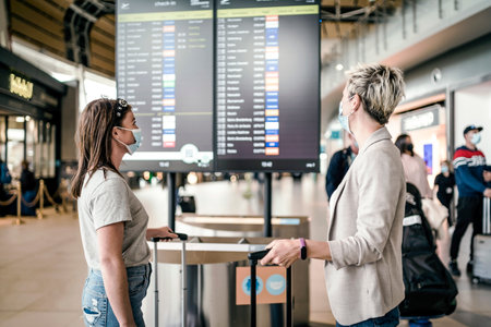 Two traveling women wearing protective masks discussing by flight information board at the Faro airportの写真素材