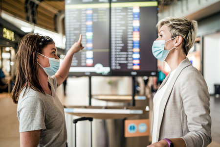 Two traveling women wearing protective masks discussing by flight information board at the Faro airportの写真素材