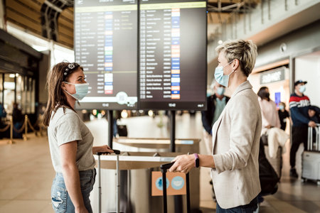 Two traveling women wearing protective masks discussing by flight information board at the Faro airportの写真素材
