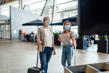 Two traveling women wearing protective masks discussing by flight information board at the Faro airportの写真素材