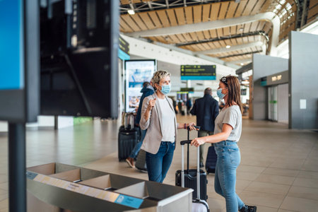 Two traveling women wearing protective masks discussing by flight information board at the Faro airportの写真素材