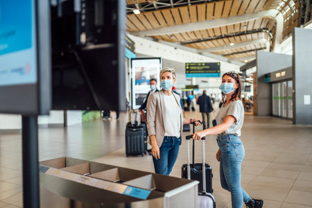 Two traveling women wearing protective masks discussing by flight information board at the Faro airportの写真素材