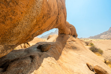 Granite peaks and rugged desert landscape at Spitzkoppe Community Campsite, Namibia. Golden grassland with scattered acacia trees contrasts against dramatic rocky mountains under a clear sky.の写真素材