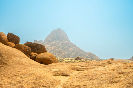 Granite peaks and rugged desert landscape at Spitzkoppe, Namibia. Golden grassland with scattered acacia trees contrasts against dramatic rocky mountains under a clear sky.の写真素材