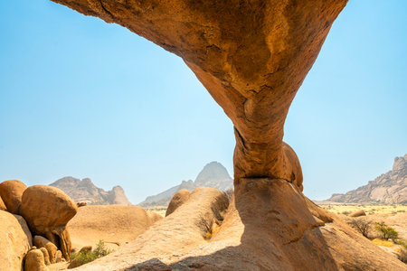Granite peaks and rugged desert landscape at Spitzkoppe, Namibia. Golden grassland with scattered acacia trees contrasts against dramatic rocky mountains under a clear sky.の写真素材