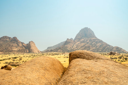Granite peaks and rugged desert landscape at Spitzkoppe Community Campsite, Namibia. Golden grassland with scattered acacia trees contrasts against dramatic rocky mountains under a clear sky.の写真素材