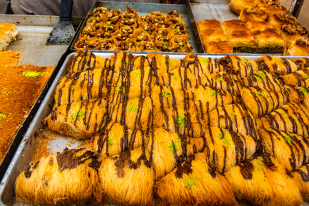 Close-up view of assorted traditional Middle Eastern pastries, including baklava and kataifi nests filled with nuts, displayed in a street food stall in Casablanca, Morocco. A vendor's hand is seen cutting pieces of the golden, syrup-glazed desserts, showcasing authentic Moroccan and Mediterranean sweetsの写真素材